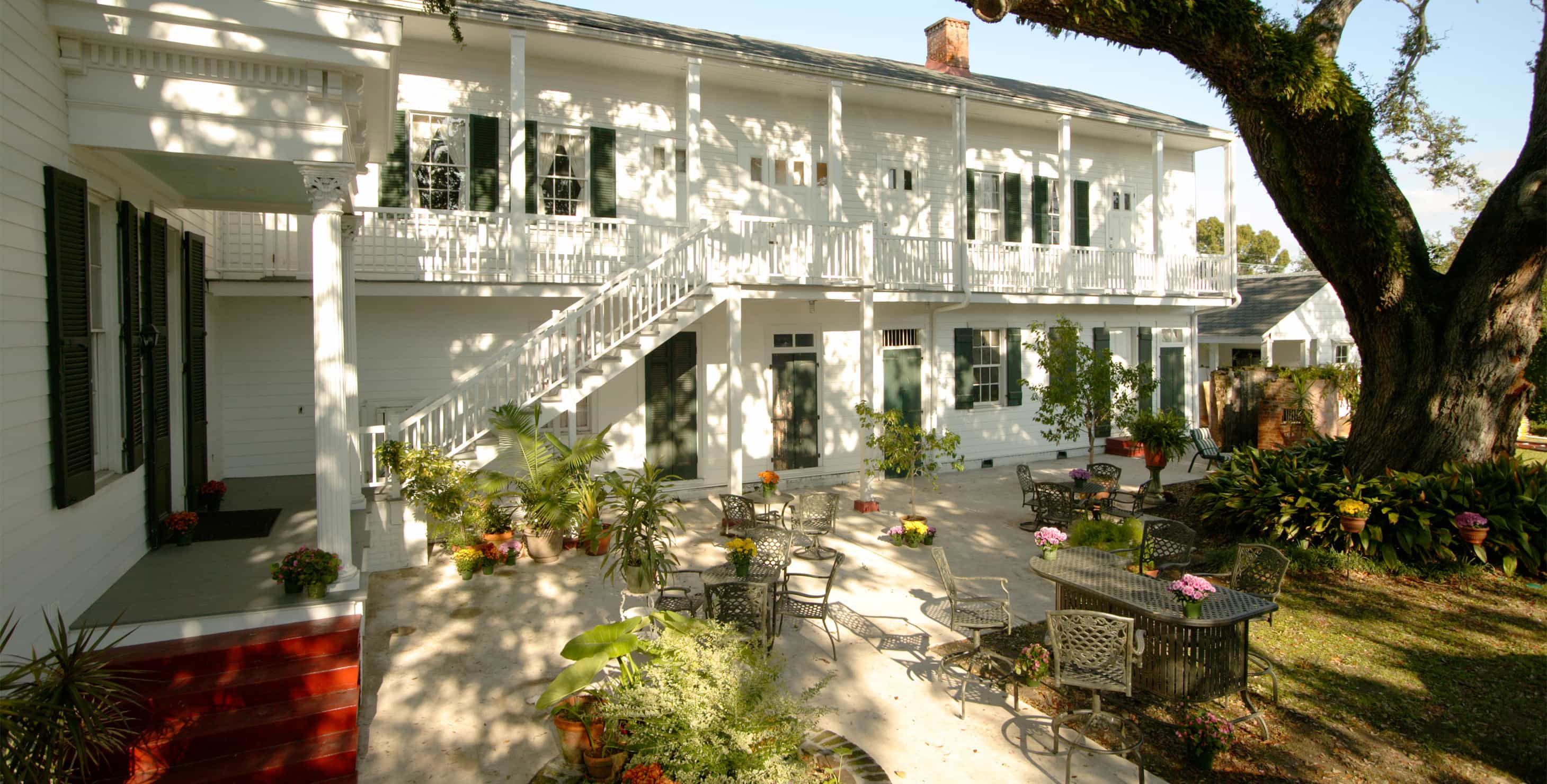 A white two-story building with a balcony and stairs leading to a patio area with tables and chairs.