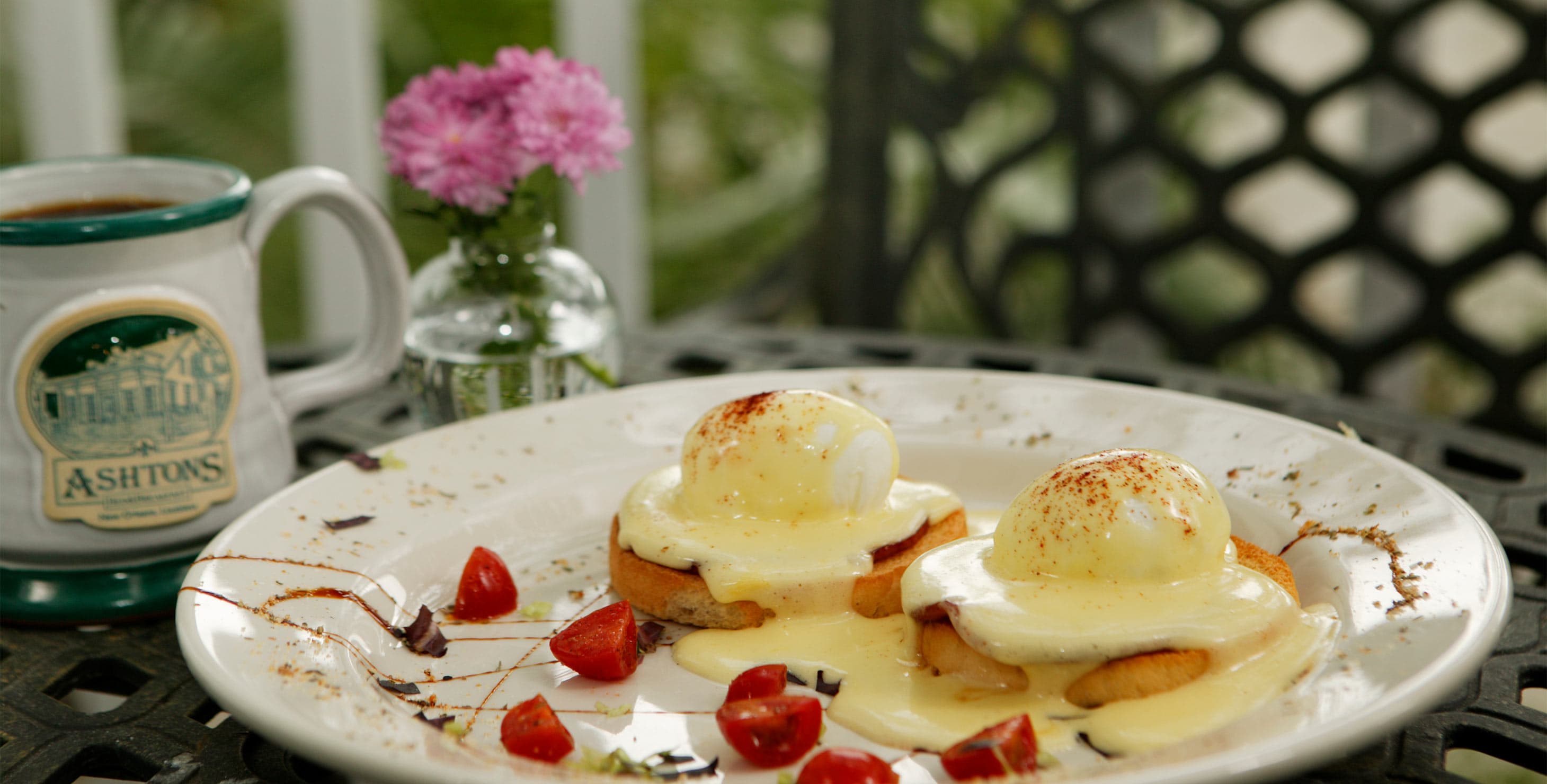 a plate of eggs benedict with strawberries and a coffee mug on a table with flowers